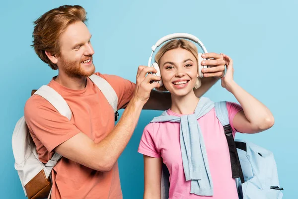 Estudiante pelirroja con mochila tocando auriculares inalámbricos de amigo rubio en azul - foto de stock