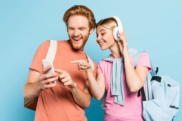 Blonde woman listening music in wireless headphones while pointing with finger at smartphone in hands of student on blue — Stock Photo