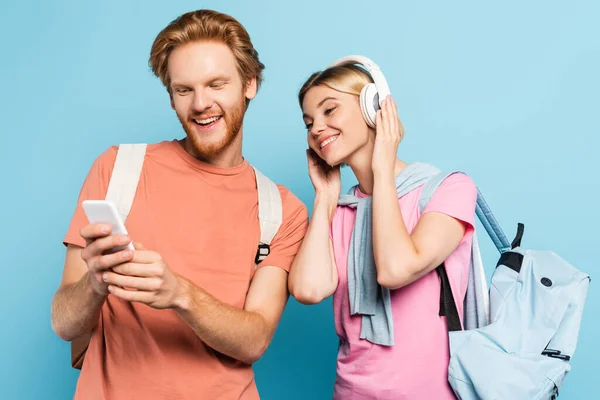 Blonde woman listening music in wireless headphones while looking at smartphone in hands of student on blue — Stock Photo