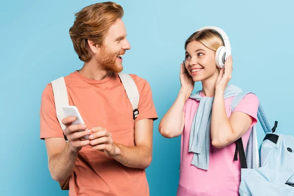 Femme blonde écouter de la musique dans les écouteurs sans fil tout en regardant l'étudiant avec smartphone sur bleu — Photo de stock