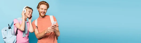 Imagen horizontal de mujer rubia escuchando música en auriculares inalámbricos mientras mira el teléfono inteligente en manos de un estudiante en azul - foto de stock