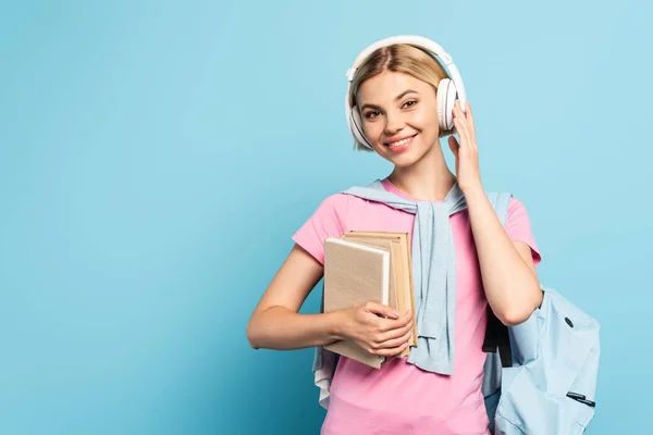 Jeune femme blonde écouter de la musique dans des écouteurs sans fil tout en tenant des livres sur bleu — Photo de stock