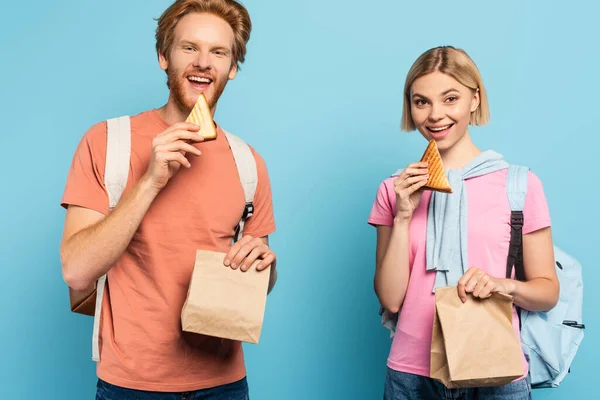 Jóvenes estudiantes sosteniendo bolsas de papel y comiendo pan tostado en azul - foto de stock