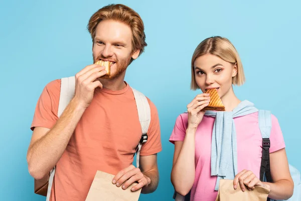 Jeunes étudiants tenant des sacs en papier et mangeant des sandwichs sur bleu — Photo de stock