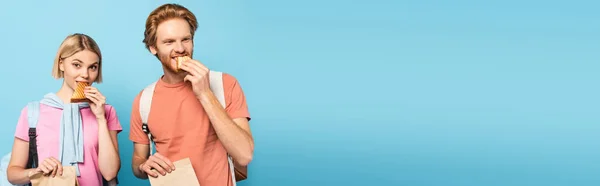 Panoramic shot of young students holding paper bags and eating sandwiches on blue — Stock Photo