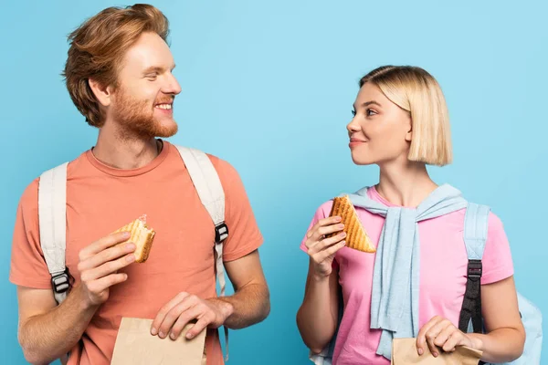 Young students holding paper bags and sandwiches while looking at each other on blue — Stock Photo