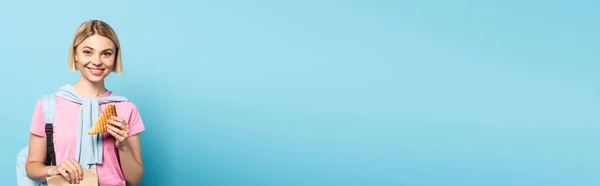 Panoramic crop of young blonde student holding paper bag and toast bread on blue — Stock Photo