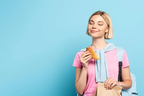 Jeune étudiante blonde les yeux fermés tenant sac en papier et pain grillé sur bleu — Photo de stock