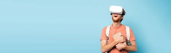 Récolte panoramique de rousse excitée étudiant en réalité virtuelle casque tenant des livres et des gestes sur bleu — Photo de stock
