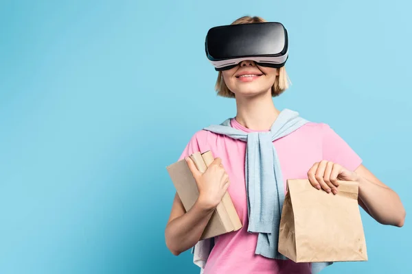 Joven estudiante rubia en auriculares de realidad virtual sosteniendo libros y bolsa de papel en azul - foto de stock