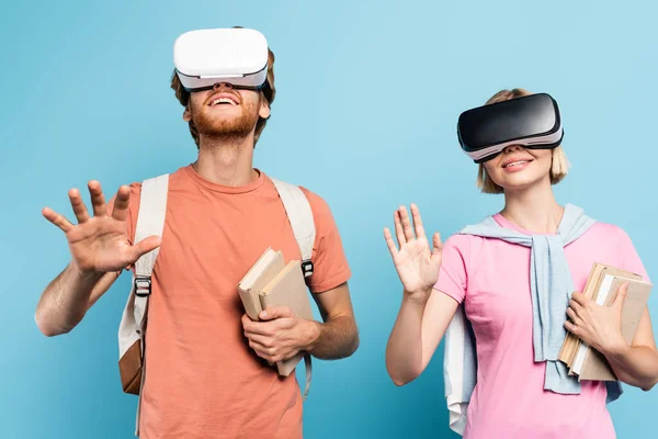 Giovani studenti in cuffie realtà virtuale in possesso di libri e gesti sul blu — Foto stock