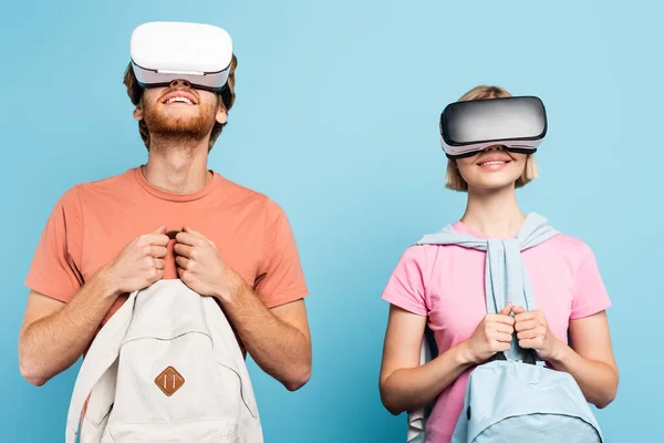 Young students in virtual reality headsets holding backpacks on blue — Stock Photo