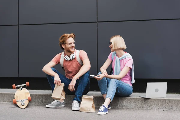 Students in glasses sitting near skateboard and gadgets while looking at each other outside — Stock Photo