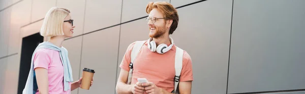 Panoramic crop of blonde student holding paper cup while looking at redhead friend with smartphone — Stock Photo