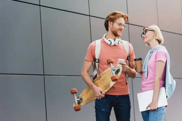 Blonde student holding paper cup and looking at friend with skateboard and smartphone standing outside — Stock Photo