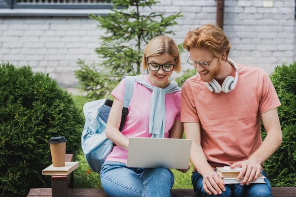 Étudiants en lunettes regardant un ordinateur portable assis sur un banc près de livres et tasse en papier — Photo de stock