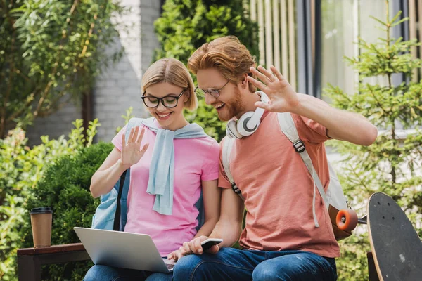 Students in glasses looking at laptop while waving hands and sitting on bench — Stock Photo