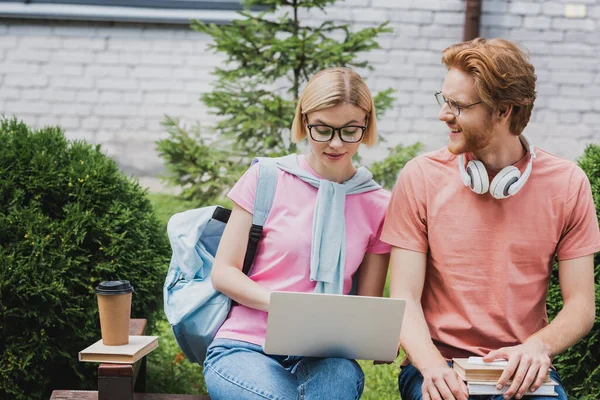 Students in glasses using laptop while sitting on bench near books and paper cup — Stock Photo