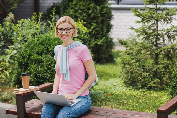 Étudiant blonde dans des lunettes à l'aide d'un ordinateur portable près de tasse en papier tout en étant assis sur le banc — Photo de stock