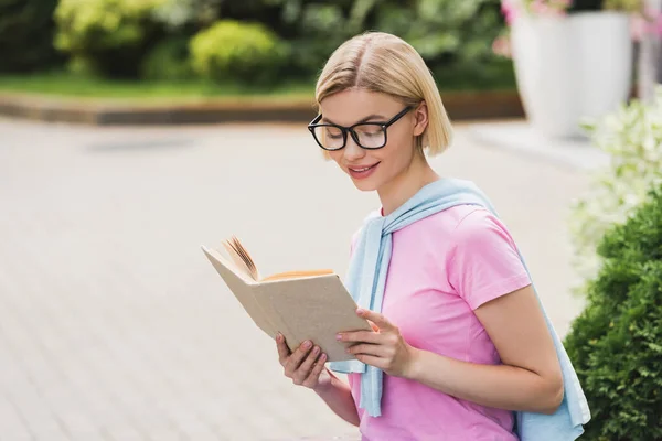 Jeune étudiante blonde dans des lunettes de lecture livre à l'extérieur — Photo de stock