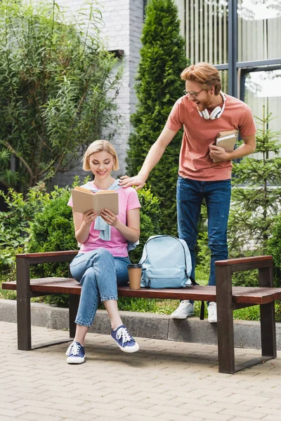Pelirroja estudiante tocando rubia amigo lectura libro mientras sentado en banco - foto de stock