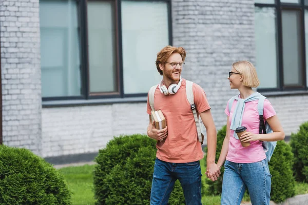 Young students in glasses holding hands and looking at each other while walking near campus — Stock Photo