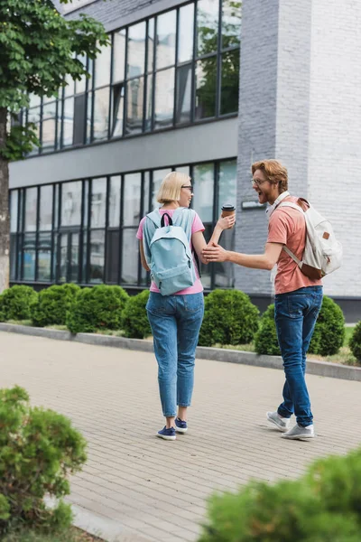 Enfoque selectivo de estudiante emocionado y barbudo tocando amigo rubio con taza de papel cerca del campus - foto de stock