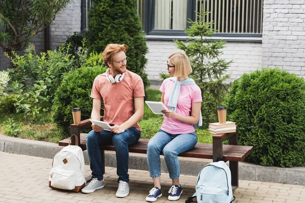 Bearded and blonde students sitting on bench near books, paper cups and backpacks while holding digital tablets — Stock Photo