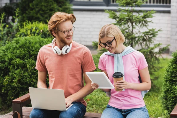 Bearded student with laptop looking at blonde friend using laptop and holding paper cup — Stock Photo