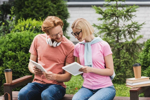 Blonde and redhead students looking at digital tablet near paper cups and books on bench — Stock Photo