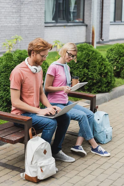 Молодые студенты, сидящие на скамейке и с помощью гаджетов во время онлайн-обучения за пределами — стоковое фото