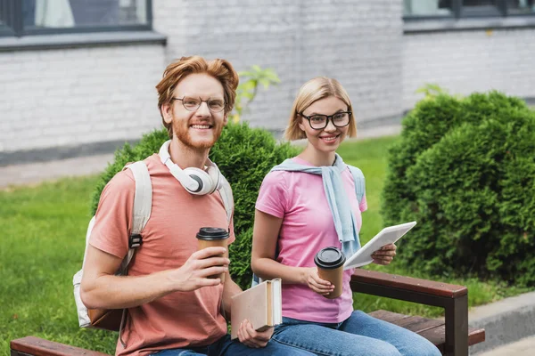 Jovens estudantes em copos segurando copos de papel, tablet digital e livros enquanto sentado no banco — Fotografia de Stock