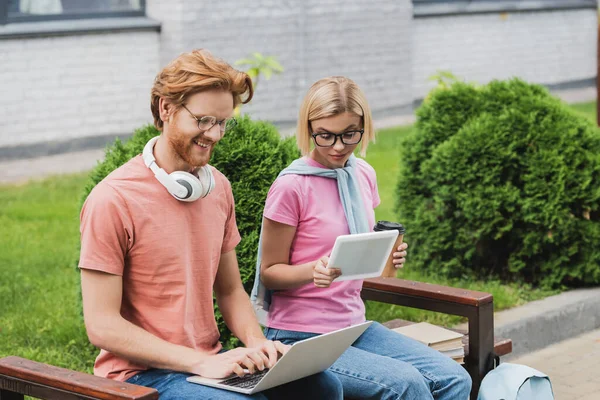 Estudiantes en gafas sentados en el banco y el uso de gadgets mientras que el estudio en línea fuera - foto de stock