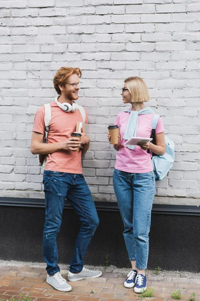 Estudantes em copos segurando café para ir e olhando uns para os outros, enquanto de pé perto da parede de tijolo — Fotografia de Stock