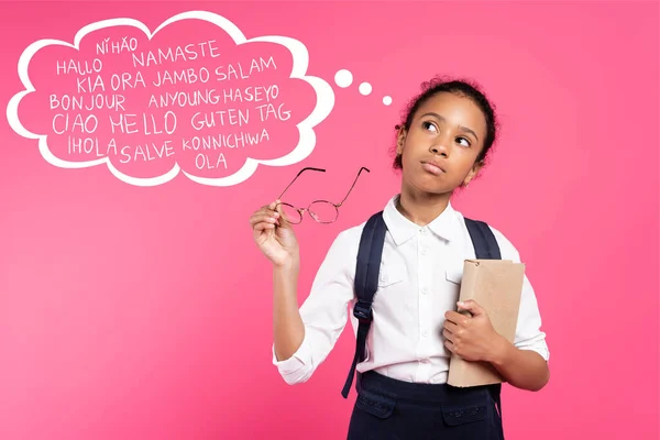 Pensive african american schoolgirl with book and eyeglasses standing near thought bubble with greeting lettering on pink — Stock Photo