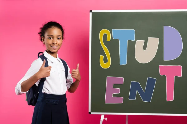 Colegiala afroamericana de pie cerca de pizarra con letras de estudiante y mostrando los pulgares hacia arriba en rosa - foto de stock