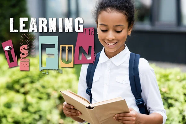 African american schoolgirl reading book near learning is fun lettering outdoors — Stock Photo