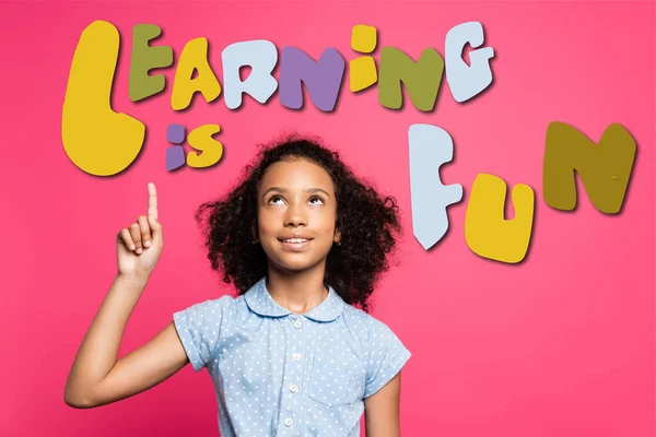 Rizado africano americano niño apuntando con el dedo y mirando hacia arriba cerca de aprender es divertido letras en rosa - foto de stock