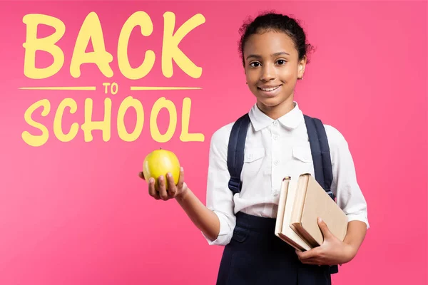 Afro-américaine écolière avec sac à dos et pomme près de retour à l'école lettrage sur rose — Photo de stock