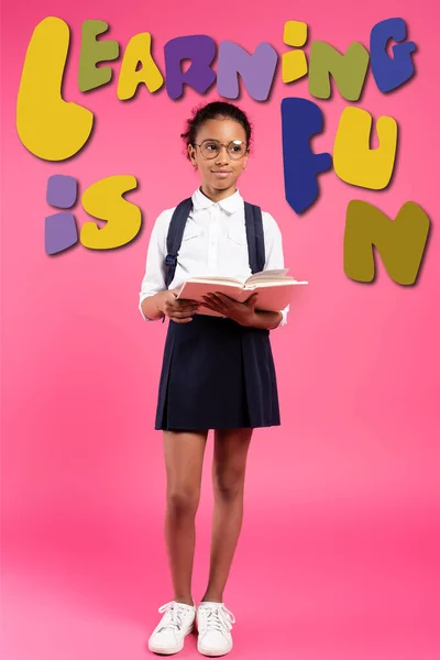 African american schoolgirl in glasses reading book near learning is fun lettering on pink — Stock Photo