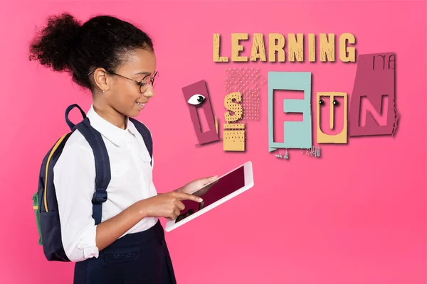 African american schoolgirl in glasses with backpack using digital tablet near learning is fun lettering on pink — Stock Photo