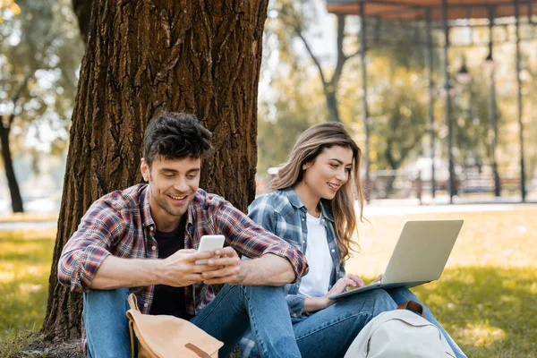Alegre pareja usando gadgets mientras sentado bajo árbol tronco - foto de stock