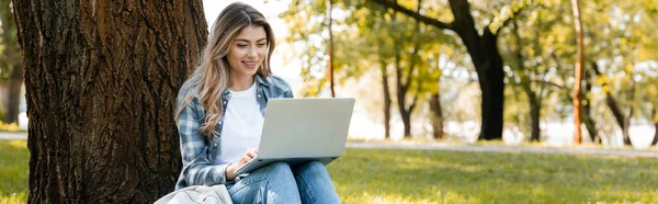 Conceito panorâmico de mulher usando laptop enquanto sentado sob tronco de árvore — Fotografia de Stock