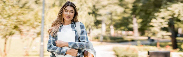 Plano panorámico de mujer en camisa a cuadros sosteniendo portátil y mirando a la cámara en el parque - foto de stock