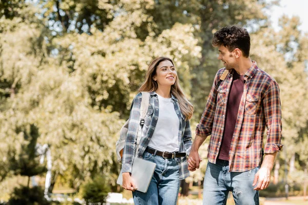 Mujer en camisa a cuadros con portátil cogido de la mano con el hombre en el parque - foto de stock