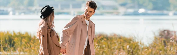 Panoramic shot of man in trench coat holding hands with woman in hat — Stock Photo