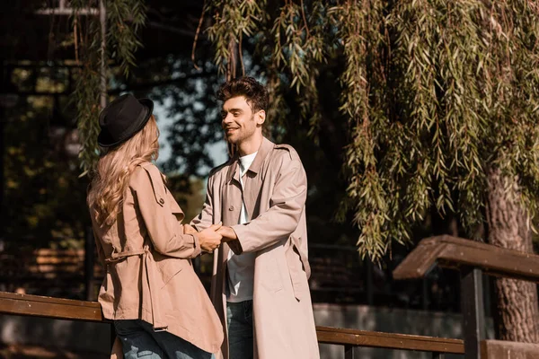 Мужчина в плаще смотрит на женщину в шляпе, держась за руки в парке — стоковое фото