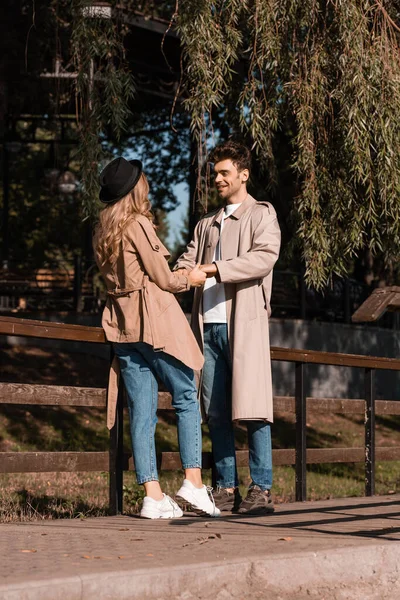 Jubelnder Mann im Trenchcoat blickt Frau mit Hut an und hält Händchen im Park — Stockfoto