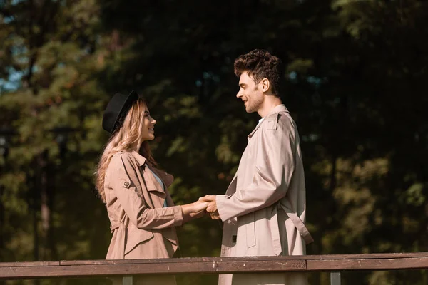 Side view of stylish couple in trench coats holding hands in park — Stock Photo