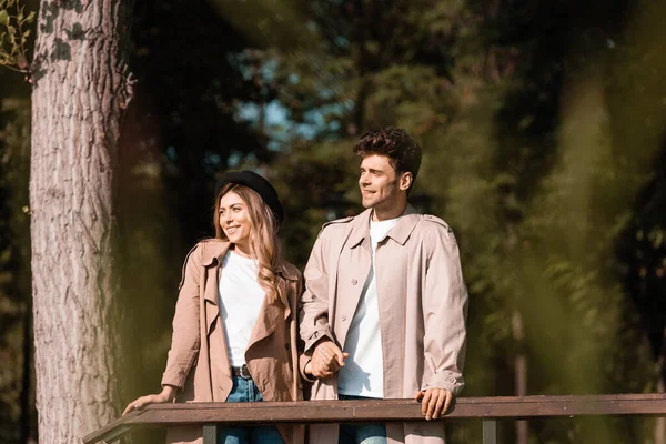 Stylish couple in trench coats looking away and holding hands in park — Stock Photo